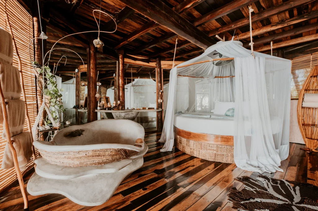 Interior of Azulik Tulum villa featuring handcrafted wooden furniture, woven fiber bed canopy netting, and stone bathtub — lit entirely by candlelight