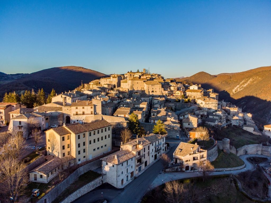 Aerial shot of a medieval Italian village with stone buildings and hills under a clear blue sky.